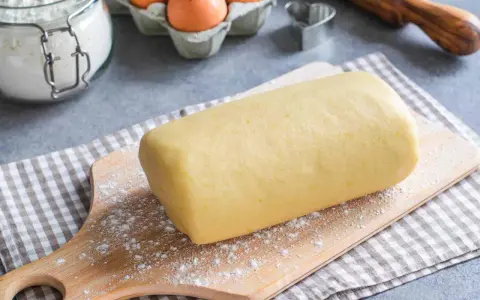 Rotolo di pasta frolla con strutto, pronto per essere trasformato in deliziosi biscotti. Ingredienti base visibili: farina, uova.