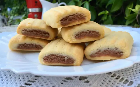 Biscotti nascondini impilati su un piatto bianco, con un ripieno scuro visibile. Sullo sfondo, una moka rossa e foglie verdi.