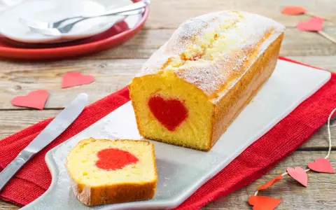 Plumcake con cuore rosso al centro, cosparso di zucchero a velo. Una fetta tagliata rivela la sorpresa. Decorazioni a forma di cuore completano l'atmosfera romantica.