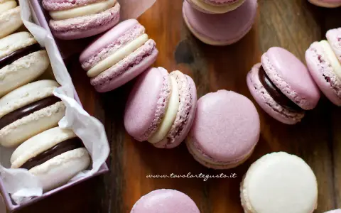 Deliziosi macaron dolci rosa e bianchi, alcuni con ripieno al cioccolato, sparsi su un tagliere di legno.