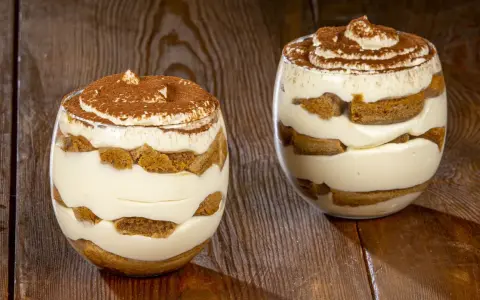 Due bicchieri di tiramisù senza uova benedetta fatto in casa, stratificato con crema e savoiardi, spolverato di cacao.