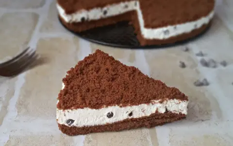 Una fetta di torta al cioccolato con ripieno cremoso al mascarpone e biscotti sbriciolati, decorata con gocce di cioccolato.