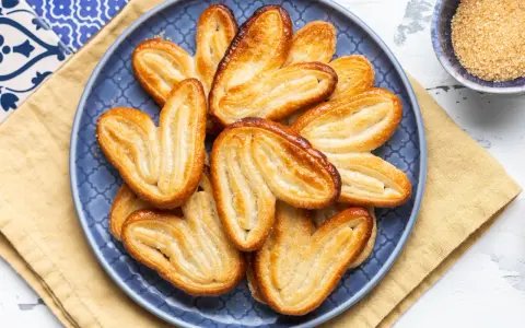 Un piatto blu pieno di croccanti ventagli di pasta sfoglia, dorati e zuccherati, pronti per essere gustati.