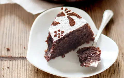 Una fetta di torta caprese ricetta facile, cosparsa di zucchero a velo e cacao, su un piatto bianco a forma di cuore.