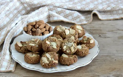 Biscotti senza zucchero, dorati e croccanti, guarniti con mandorle a lamelle. Accanto, una ciotola piena di mandorle intere.