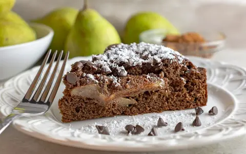 Una fetta di torta pere e cioccolato, cosparsa di zucchero a velo e gocce di cioccolato, con pere caramellate all'interno.