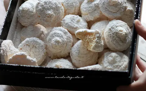 Scatola piena di soffici dolcetti al cocco, spolverati di zucchero a velo. Uno è spezzato, mostrando l'interno friabile.