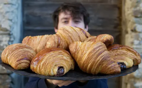 Un ragazzo sorride dietro un vassoio pieno di croissant brioche dorati e fragranti, alcuni con ripieno di cioccolato.
