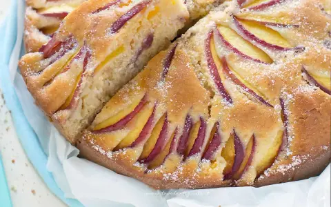 Una deliziosa torta di pesche e yogurt greco, con fette di pesca disposte a raggiera sulla superficie dorata e cosparsa di zucchero a velo.