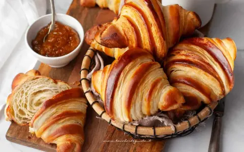 Croissant dorati e sfogliati, un invito a scoprire la loro origine. Marmellata in ciotola e una tazza bianca completano la scena.