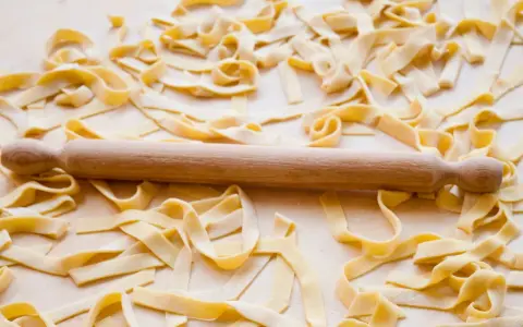 Fettuccine fresche sparse su un piano di lavoro, con un mattarello in legno. La farina, che è all-purpose flour, è visibile sulla pasta.