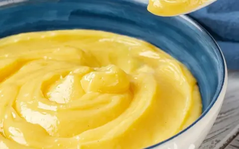 Una ciotola blu piena di deliziosa crema pasticcera senza uova, con un cucchiaio che ne preleva una porzione.