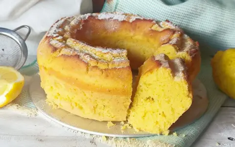 Torta soffice e dorata, spolverata di zucchero a velo, preparata con dolci con eritritolo. Accanto, limoni freschi e un setaccio.