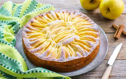 Torta di mele con yogurt, decorata con fettine di mela e zucchero a velo, su un piatto grigio, accanto a un tovagliolo a righe verdi e bianche.