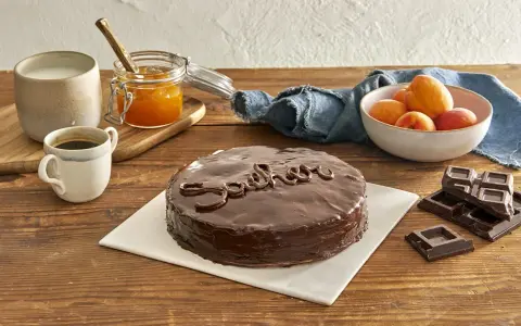 Torta Sacher, un classico viennese, con la sua glassa lucida e il nome scritto in cioccolato. Accanto, albicocche, marmellata e caffè, ingredienti che evocano le sue origini.