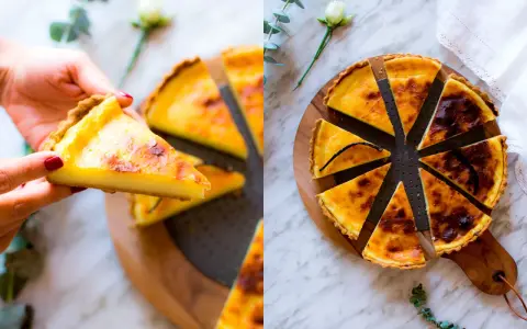 Una mano prende una fetta di flan parisien dorato. La torta intera, tagliata a spicchi, è su un tagliere di legno, decorata con un fiore e un rametto di eucalipto.