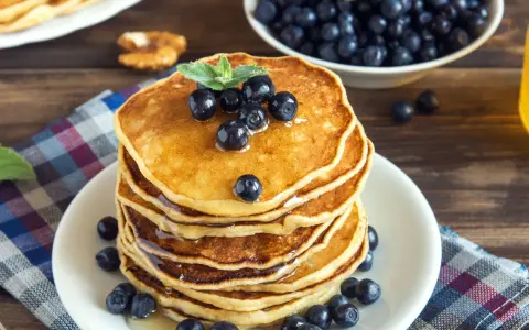 Pila di pancake vegani dorati, guarniti con mirtilli freschi e sciroppo d'acero, su un piatto bianco, con una ciotola di mirtilli e una bottiglia di sciroppo sullo sfondo.