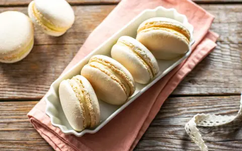 Macarons bianchi e dorati, perfetti esempi di come sono fatti i macarons, con ripieno cremoso, su un piatto bianco e tovagliolo rosa.
