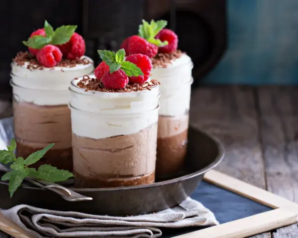 Tre deliziosi bicchierini di dolce freddo al cioccolato e panna, guarniti con lamponi freschi e scaglie di cioccolato.