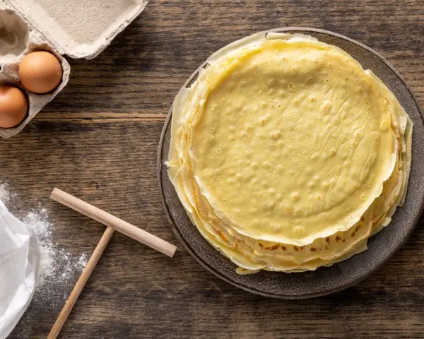 Pila di crepes dorate, pronte per essere farcite. Accanto, uova fresche e farina, ingredienti perfetti per crepes 2 uova.