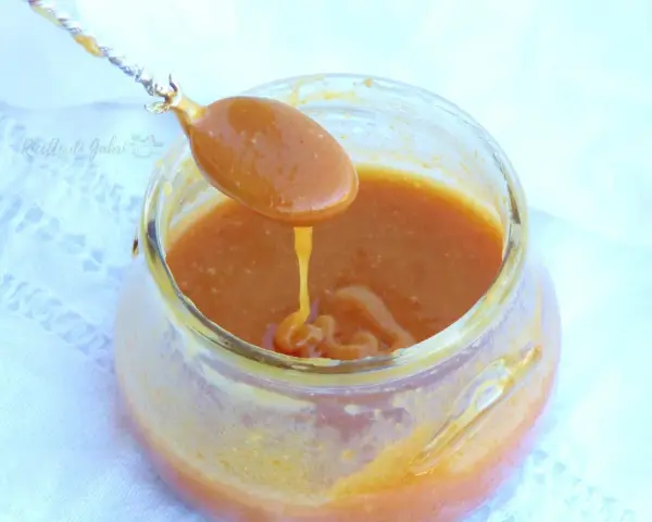 Un cucchiaio raccoglie un denso caramello salato da un barattolo di vetro. La salsa dorata cola lentamente, invitando a un assaggio goloso.
