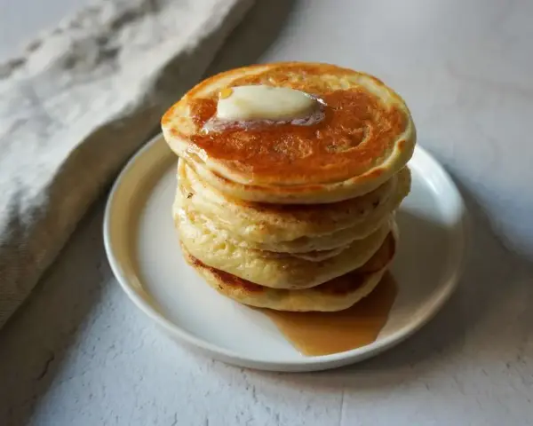 Pancakes dorati e soffici, impilati su un piatto bianco, con una noce di burro che si scioglie in cima e sciroppo d'acero che cola. Sembrano pronti per essere gustati, un vero piacere per gli occhi e per il palato, come un pancake in bottiglia pronto d...