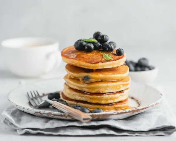 Pancake con farina di riso, guarniti con mirtilli freschi e sciroppo d'acero, su un piatto rustico con forchetta.