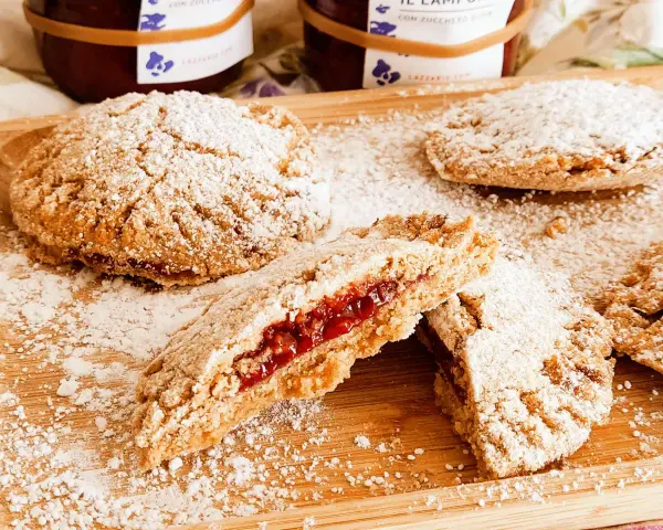 Biscotti integrali con marmellata, spolverati di zucchero a velo, su un tagliere di legno. Un morso rivela il ripieno rosso e invitante.