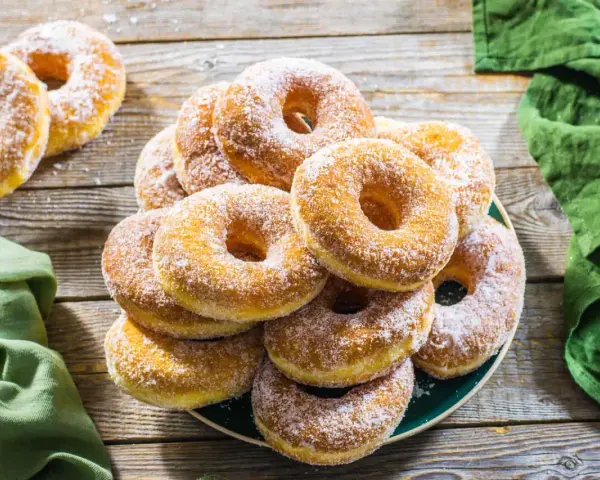 Un piatto pieno di graffe napoletane, dolci soffici e dorati, cosparse di zucchero a velo, su un tavolo di legno rustico.