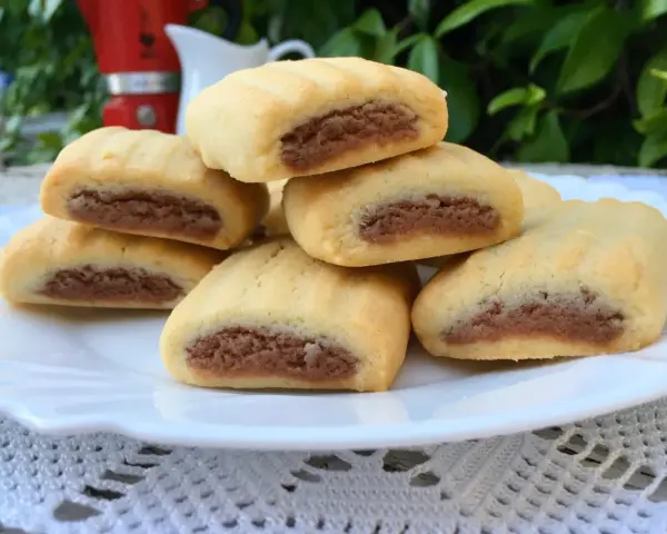 Biscotti nascondini impilati su un piatto bianco, con un ripieno scuro visibile. Sullo sfondo, una moka rossa e foglie verdi.