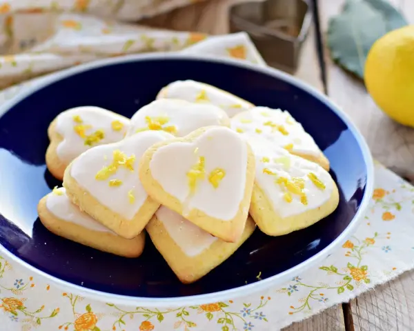 Cuore di biscotti al limone glassati, decorati con scorza di limone, su un piatto blu.