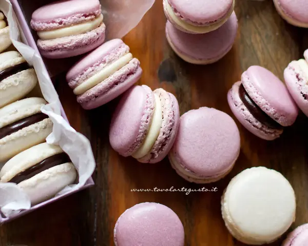 Deliziosi macaron dolci rosa e bianchi, alcuni con ripieno al cioccolato, sparsi su un tagliere di legno.