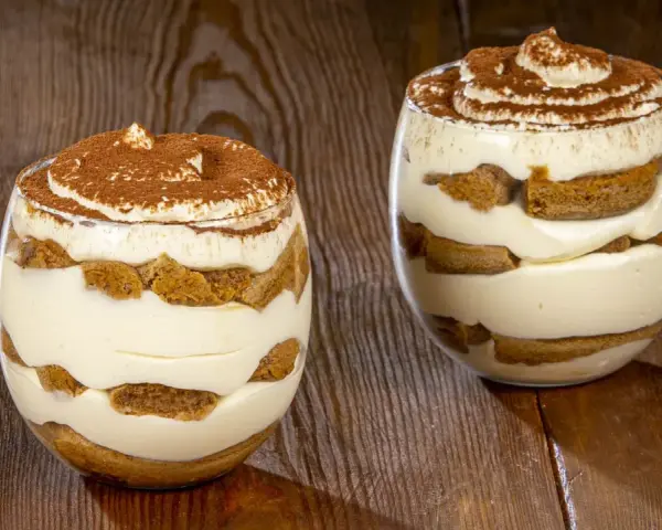 Due bicchieri di tiramisù senza uova benedetta fatto in casa, stratificato con crema e savoiardi, spolverato di cacao.