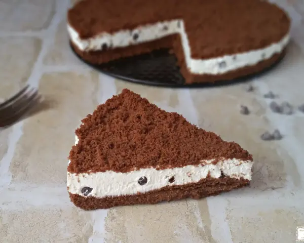 Una fetta di torta al cioccolato con ripieno cremoso al mascarpone e biscotti sbriciolati, decorata con gocce di cioccolato.