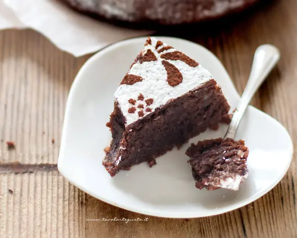 Una fetta di torta caprese ricetta facile, cosparsa di zucchero a velo e cacao, su un piatto bianco a forma di cuore.