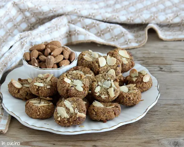 Biscotti senza zucchero, dorati e croccanti, guarniti con mandorle a lamelle. Accanto, una ciotola piena di mandorle intere.