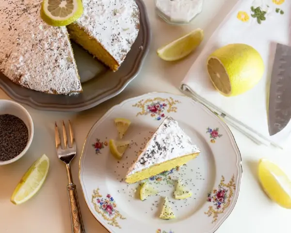Una fetta di torta al limone cosparsa di zucchero a velo e semi di papavero, guarnita con spicchi di limone fresco.