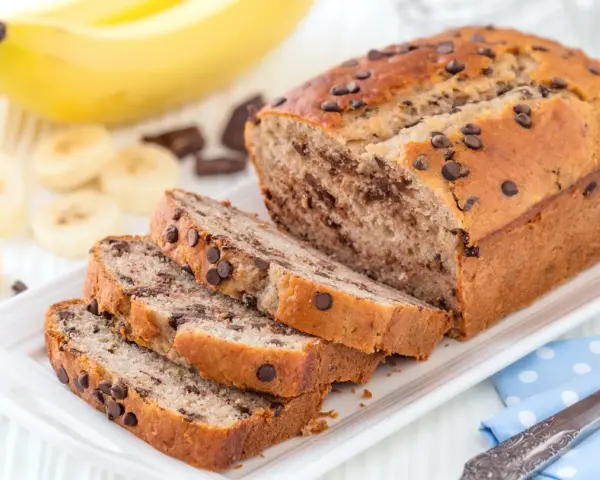 Fette di plumcake banana e cioccolato su un piatto bianco, con banane e gocce di cioccolato sullo sfondo.