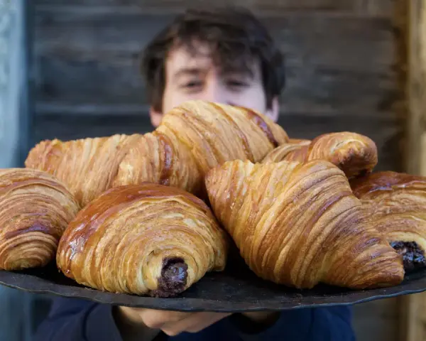 Un ragazzo sorride dietro un vassoio pieno di croissant brioche dorati e fragranti, alcuni con ripieno di cioccolato.