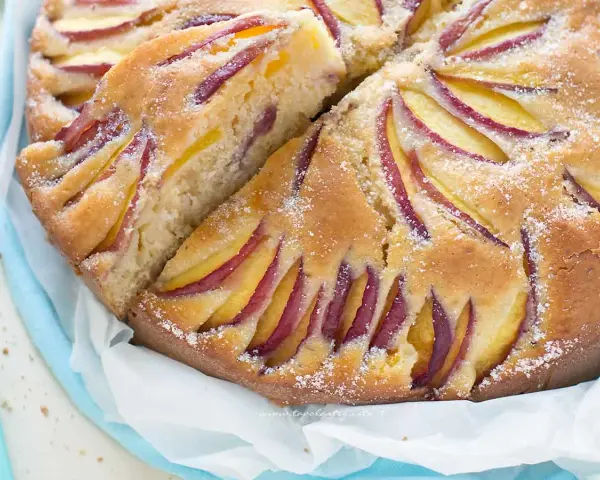 Una deliziosa torta di pesche e yogurt greco, con fette di pesca disposte a raggiera sulla superficie dorata e cosparsa di zucchero a velo.