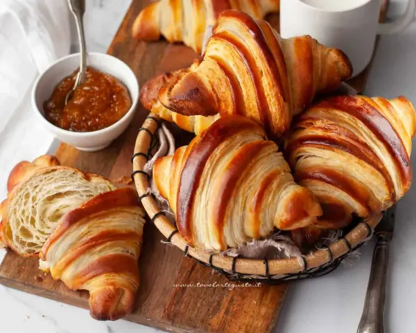 Croissant dorati e sfogliati, un invito a scoprire la loro origine. Marmellata in ciotola e una tazza bianca completano la scena.