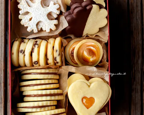 Scatola piena di biscotti al burro: a fiocco di neve, con gocce di cioccolato, ripieni di caramello, a forma di cuore e ricoperti di cioccolato.
