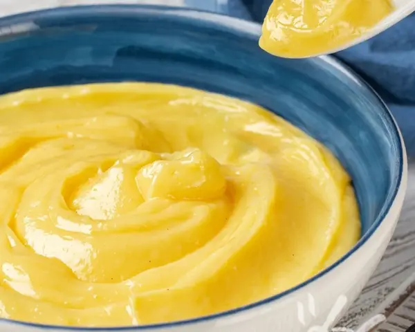Una ciotola blu piena di deliziosa crema pasticcera senza uova, con un cucchiaio che ne preleva una porzione.