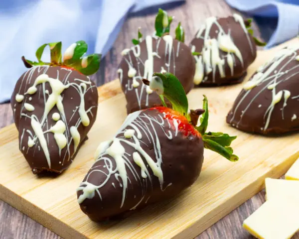 Fragole ricoperte di cioccolato fondente e decorate con cioccolato bianco. Un delizioso assaggio di frutta al cioccolato su un tagliere di legno.