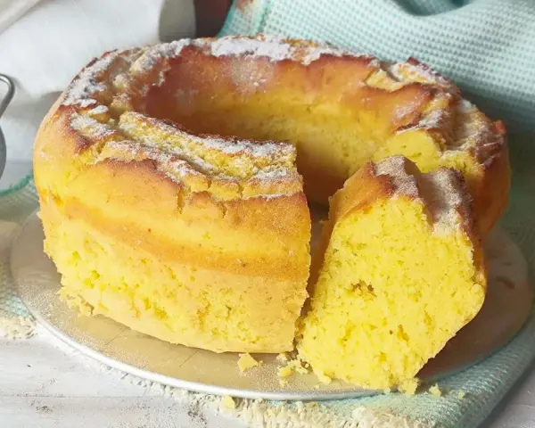 Torta soffice e dorata, spolverata di zucchero a velo, preparata con dolci con eritritolo. Accanto, limoni freschi e un setaccio.