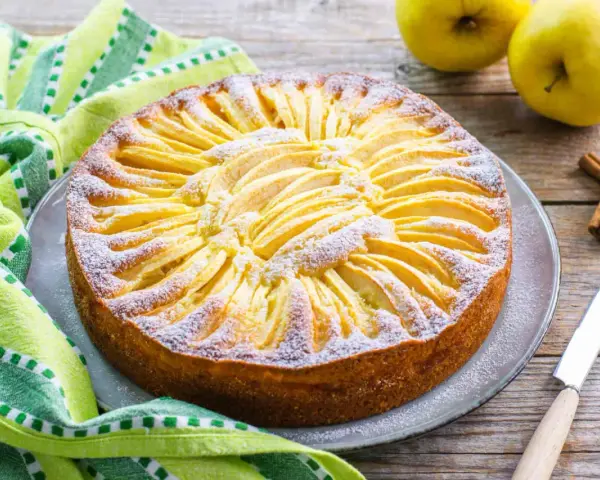 Torta di mele con yogurt, decorata con fettine di mela e zucchero a velo, su un piatto grigio, accanto a un tovagliolo a righe verdi e bianche.