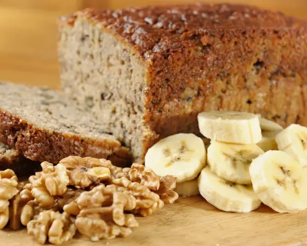 Fetta di banana bread senza uova, con noci e banane a fette, su tagliere di legno.