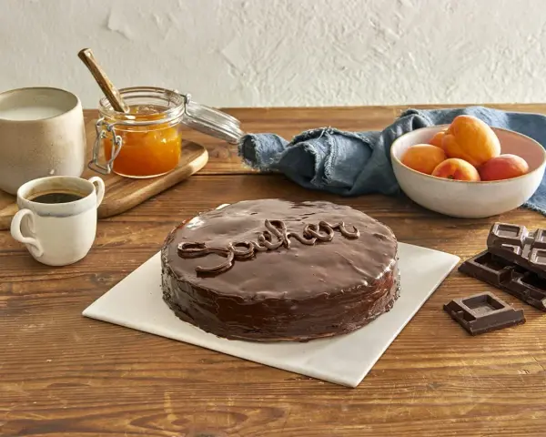 Torta Sacher, un classico viennese, con la sua glassa lucida e il nome scritto in cioccolato. Accanto, albicocche, marmellata e caffè, ingredienti che evocano le sue origini.