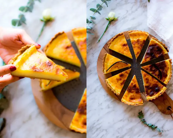 Una mano prende una fetta di flan parisien dorato. La torta intera, tagliata a spicchi, è su un tagliere di legno, decorata con un fiore e un rametto di eucalipto.