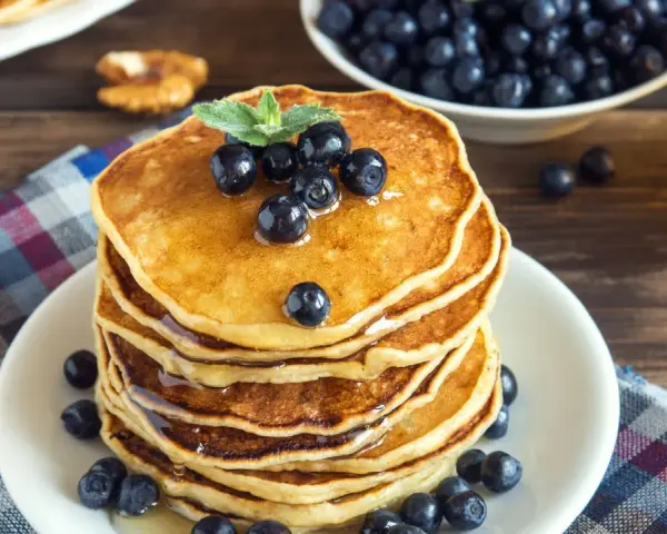 Pila di pancake vegani dorati, guarniti con mirtilli freschi e sciroppo d'acero, su un piatto bianco, con una ciotola di mirtilli e una bottiglia di sciroppo sullo sfondo.